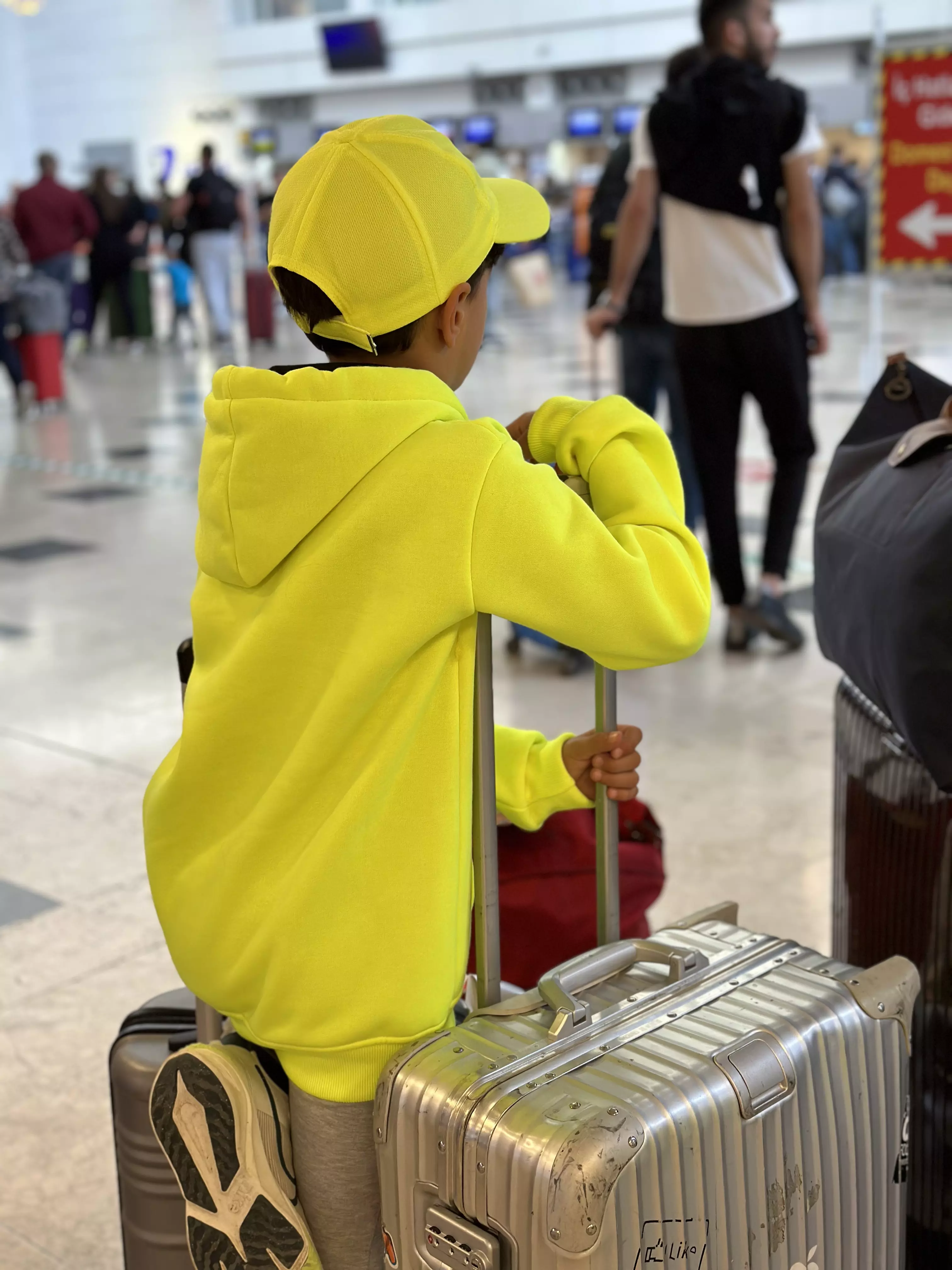 Kinder in hellen, auffälligen Outfits, besonders an belebten Orten wie Bahnhöfen oder Flughäfen, um sie leichter im Blick zu behalten und die Sicherheit zu erhöhen. Hier steht ein Junge in einem neon Kapuzenpulli neben Koffern in der Halle eines Flughafens. Durch die knallige Farbe ist er leicht zu erkennen.