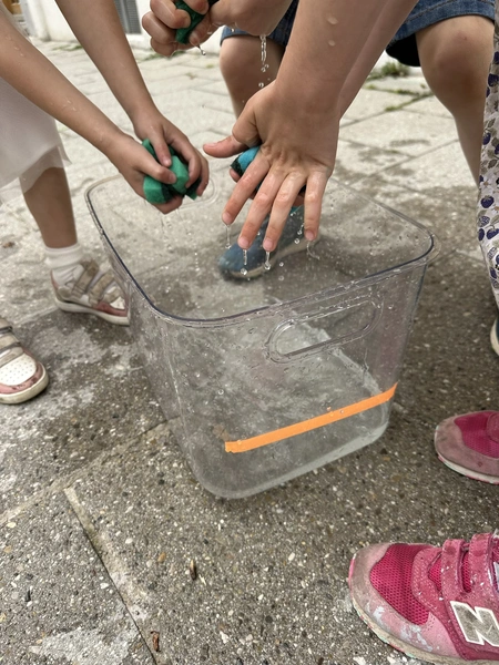 Kinder transportieren triefend nasse Schwämme von einem vollen zu einem leeren Eimer und quetschen sie aus, um den Eimer bis zu einer Markierung zu füllen. Dieses Spiel fördert Teamarbeit und macht den Kindern großen Spaß, gemeinsam eine Herausforderung zu meistern.